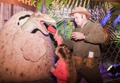 zwei Personen füttern eine lebensgroße Dinosaurierfigur, die aus einem großen Ei schaut