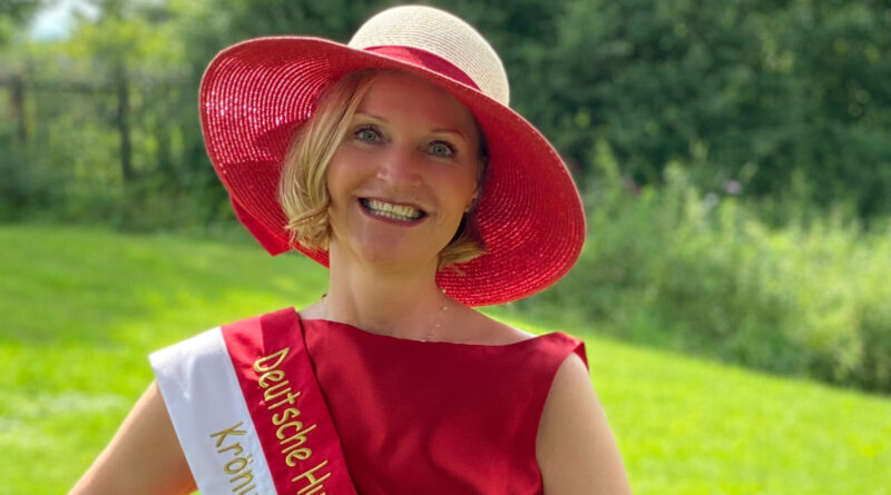 Die aktuelle Deutsche Hutkönigin Sabine B. Berner mit rotem Strohhut, rotem Kleid und einer Schärpe steht auf einer Wiese
