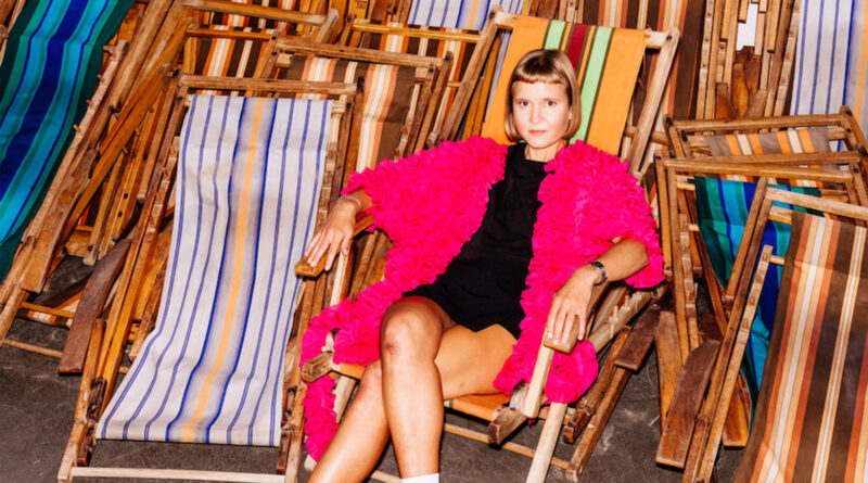 Die Autorin Caroline Wahl sitzt in einem pinken Mantel inmitten aufgestapelter Liegestühle