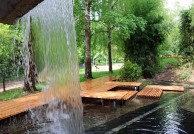 Blick durch einen kleinen Wasserfall auf das neu hergerichtete Becken des waserspielplatzes mit Holzstegen und Plattformen zum Sitzen und Erholen