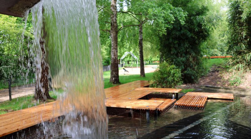 Blick durch einen kleinen Wasserfall auf das neu hergerichtete Becken des waserspielplatzes mit Holzstegen und Plattformen zum Sitzen und Erholen