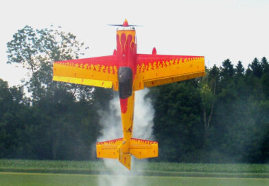 ein rotes Modelflugzeug mit gelben Flammen bemalt steigt senkrecht vor einem waldigen Hintergrund in die Luft und hinterlässt Rauchschwaden