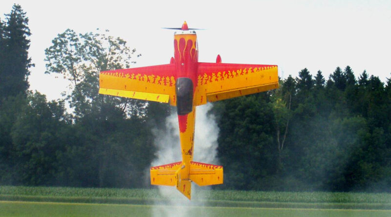 ein rotes Modelflugzeug mit gelben Flammen bemalt steigt senkrecht vor einem waldigen Hintergrund in die Luft und hinterlässt Rauchschwaden