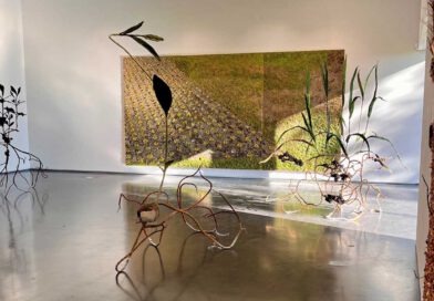 In einem Ausstellungssaal hängt ein großes Bild, das eine Beet und Rasenfläche zeigt, davor stehen große Skulpturen, die wie filigrane Pflanzen aussehen