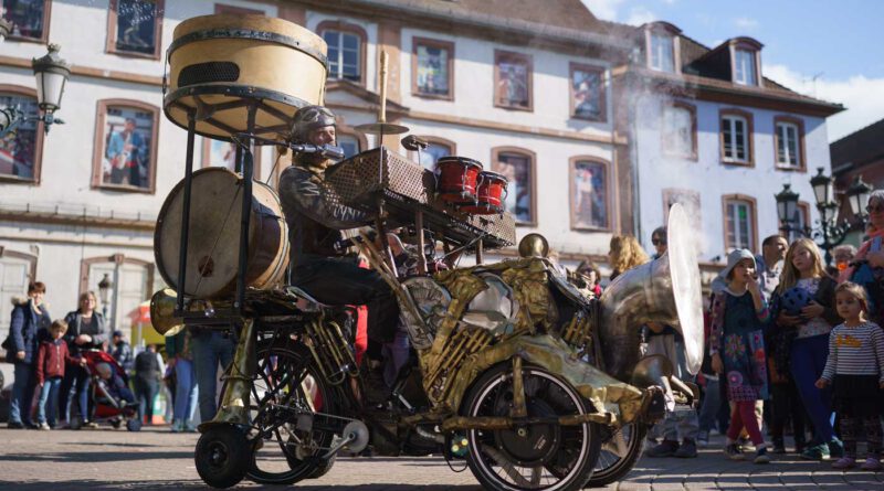 Ein Musiker sitzt auf einem Dreirad, das mit allerlei Instrumenten bestückt ist. Er ist umringt von Publikum auf einem Platz in einer Altstadt