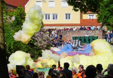 Aus großen Rohren quillt bunter Schaum und ergießt sich über das Atrium des Westertorplatzes. Eine vom Wind gelöste Schaumwolke weht durch die Luft. Das davor stehende Publikum beobachtete as Spektakel.