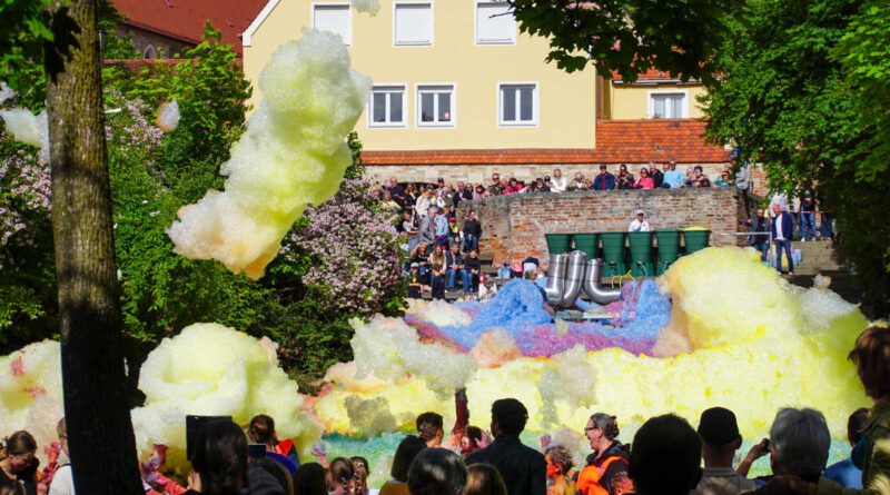 Aus großen Rohren quillt bunter Schaum und ergießt sich über das Atrium des Westertorplatzes. Eine vom Wind gelöste Schaumwolke weht durch die Luft. Das davor stehende Publikum beobachtete as Spektakel.