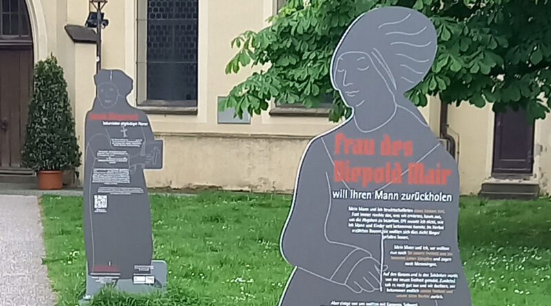 Lebensgroße Stelen, die Umrisse historischer Figuren darstellen auf denen ihre Biografie und geschichtliche Fakten gedruckt sind, stehen auf dem Rasen vor der Martinskirche