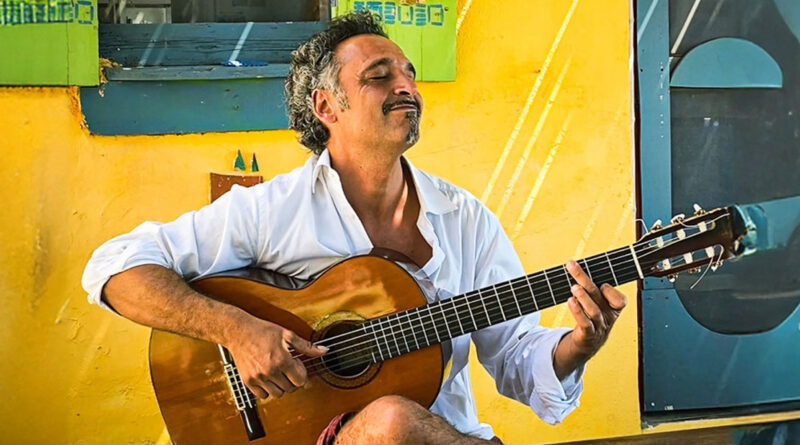 Der Gitarrist Manuel Randi sitzt mit geschlossenen Augen vor einer gelben Hauswand und spielt auf seinem Instrument