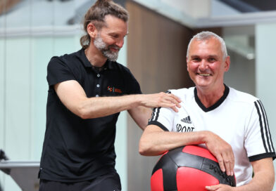 Ein Mann mit einem weißen Shirt hält einen Trainingsball in der Hand und wird von einem Rehamitarbeiter angeleitet.