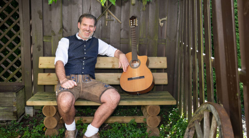 Der Kabarettist Vogelbauer in bayerischer Landhaustracht auf einer Gartenbank, neben sich eine Gitarre
