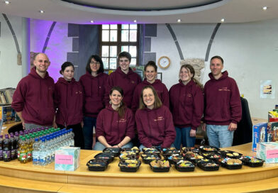 <span style="font-size: 16px;">25. Oktober, Memmingen </span><br>Stadtbibliothek lädt zum Spielen ein eine Gruppe Menschen steht hinter dem Tresen der Stadtbibliothek. Alle tragen einen weinroten Hoodie