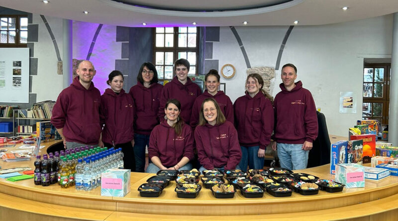 eine Gruppe Menschen steht hinter dem Tresen der Stadtbibliothek. Alle tragen einen weinroten Hoodie