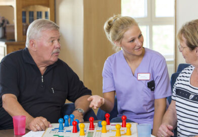Neu im AOK-Pflegenavigator: häusliche Beratungsbesuche eine Pflegekraft spielt mit zwei älteren Menschen in häuslicher Umgebung ein Brettspiel