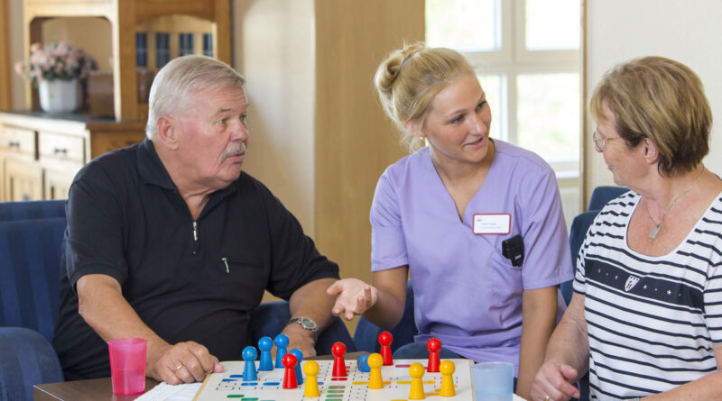 eine Pflegekraft spielt mit zwei älteren Menschen in häuslicher Umgebung ein Brettspiel