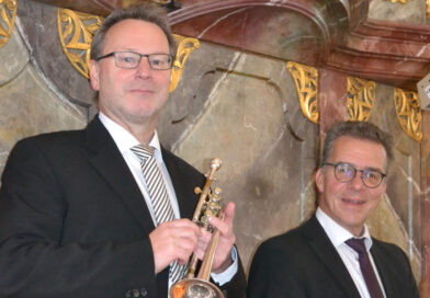 Zwei Männer in schwarzen Anzügen stehen vor einer barock verzierten Wand