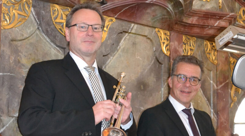 Zwei Männer in schwarzen Anzügen stehen vor einer barock verzierten Wand