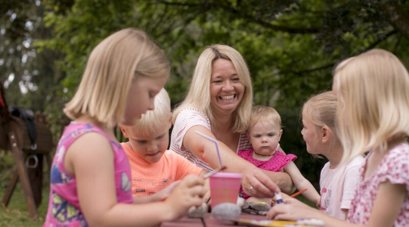 Eine lachende Frau spielt mit Kindern an einem Tisch in einer Grünanlage