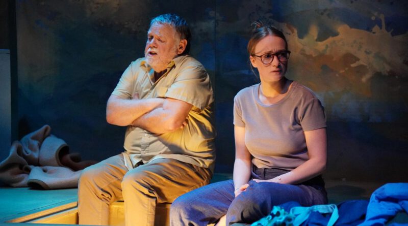 Auf einer schummerig beleuchteten Bühne sitzen ein älterer Mann und eine junge Frau mit besorgten Minen