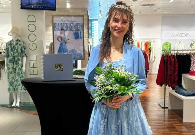 eine lächelnde junge Frau in einem hellblauen Kleid steht mit einem Blumenstrauß in der Hand in einem Modegeschäft