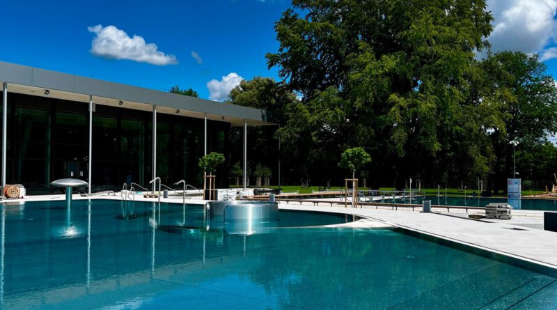 Der Außenbereich eines Schwimmbads mit einem Pool, im Hintergrund ein großer Baum und blauer Himmel