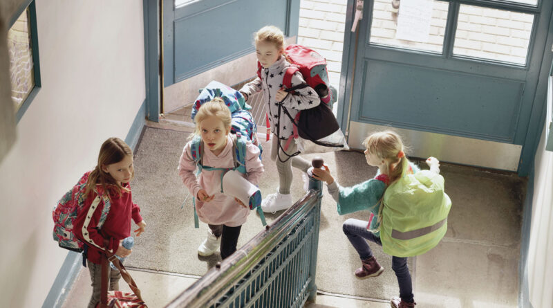 AOK-Aktion „Schulranzen Fit“ für gesunde Kinderrücken Schulkinder mit Schulranzen und Taschen gehen durch ein Treppenhaus in einem Schulgebäude.